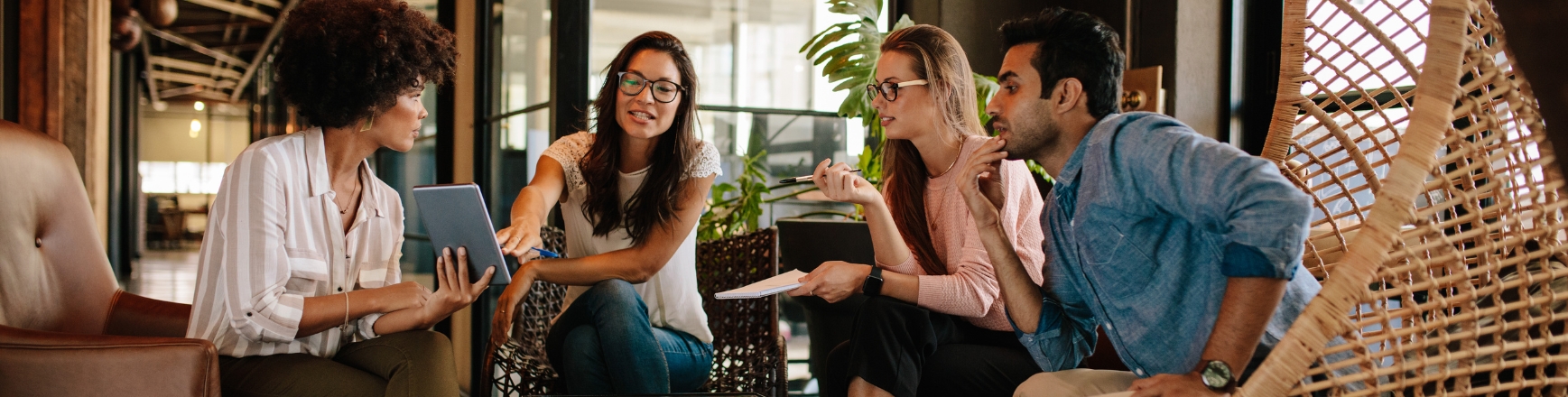 A diverse group of four colleagues sitting in a modern office lounge area, reviewing a tablet together during a corporate retreat planning discussion.
