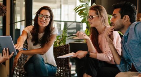 A diverse group of four colleagues sitting in a modern office lounge area, reviewing a tablet together during a corporate retreat planning discussion.