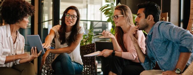A diverse group of four colleagues sitting in a modern office lounge area, reviewing a tablet together during a corporate retreat planning discussion.