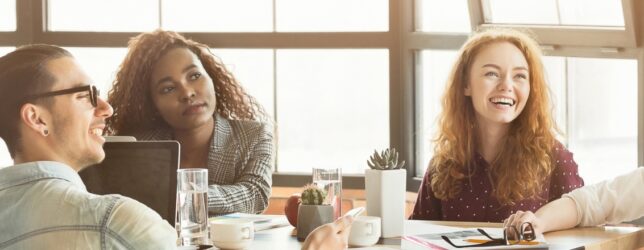 Team offsite planning discussion with a small group of colleagues gathered around a table in a bright meeting space, smiling and engaged in conversation while reviewing ideas and notes during an informal strategy session.