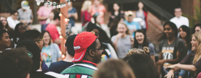 Large group of people standing in a circle outdoors, talking and laughing during a company retreat activity.